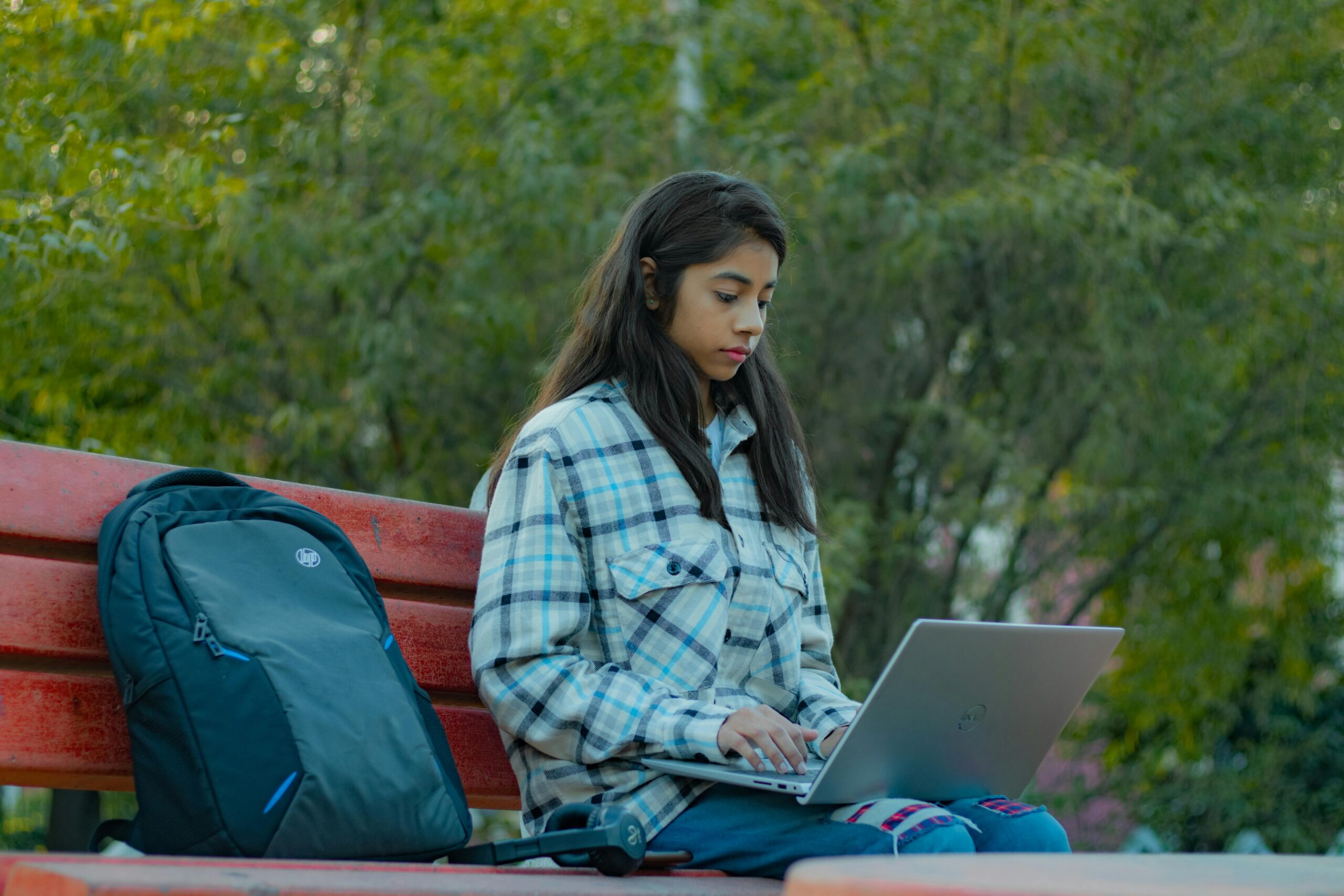 South Asian woman in plaid shirt using laptop on bench in park setting.